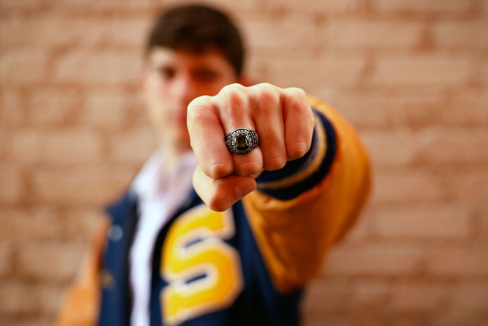 Young man in varsity jacket showing championship ring on fist toward camera against brick wall.