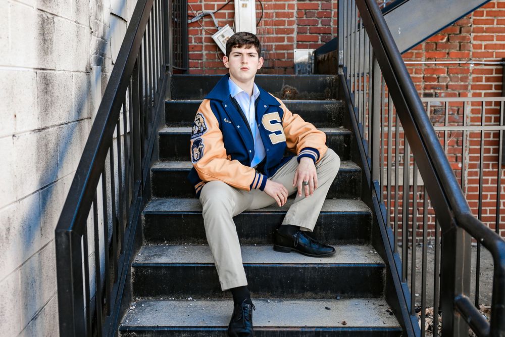 Young man in blue varsity jacket sits on metal stairs outdoors for senior portrait photo.