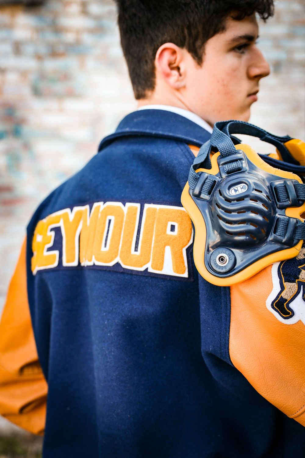Teen boy wearing a navy and gold Seymour varsity letterman jacket holding a catcher's mask outdoors.