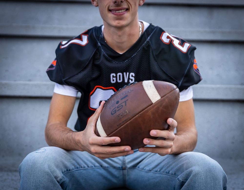 Young football player in black GOVS jersey holding a football, sitting on bleachers in jeans.
