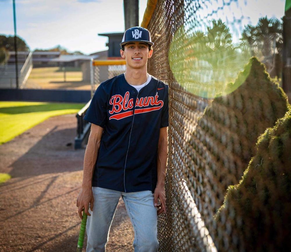 Young baseball player in Blount jersey and cap leans against chain-link fence at baseball field.