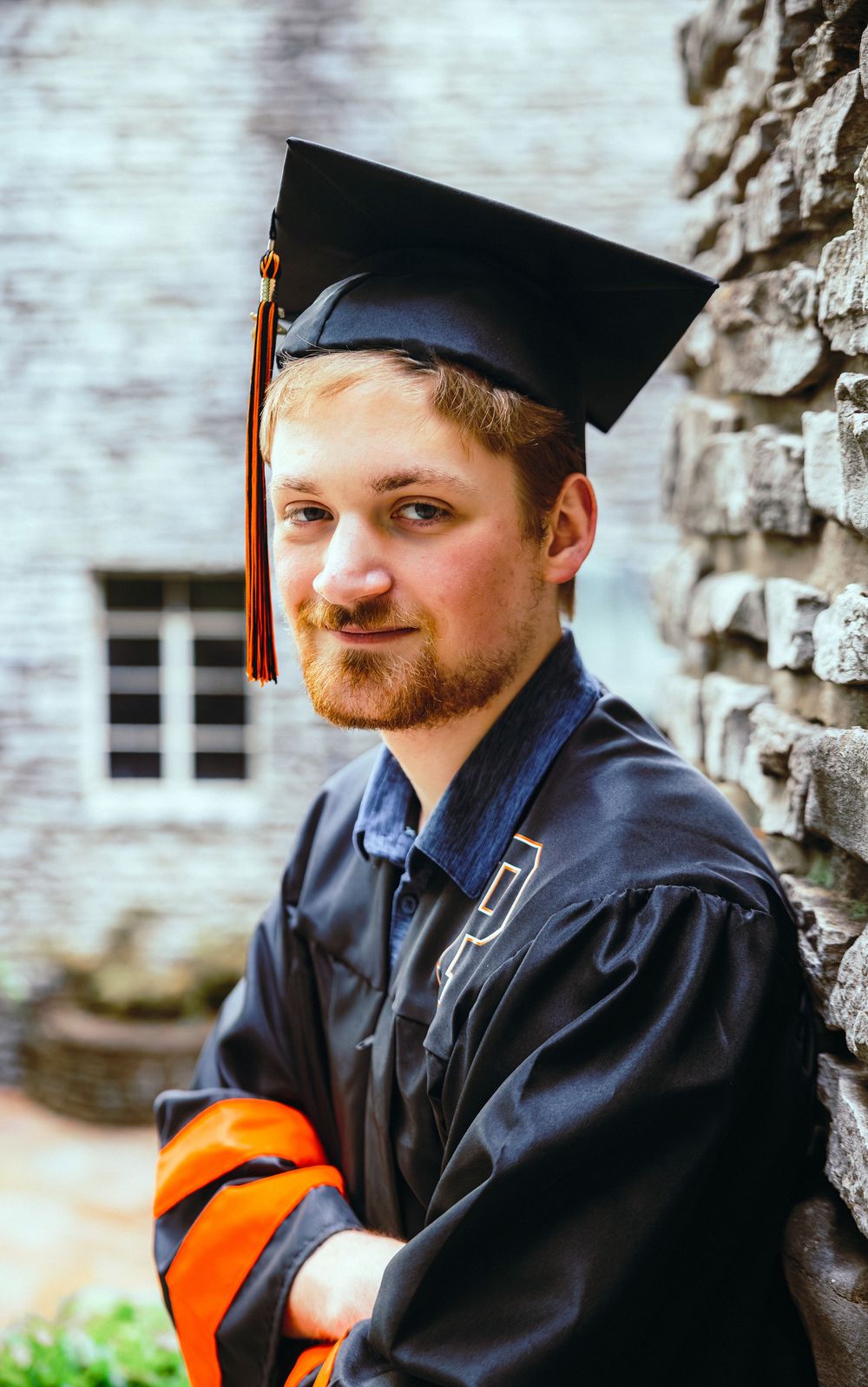 Young man in black graduation cap and gown with orange hood, leaning against stone wall, smiling confidently.
