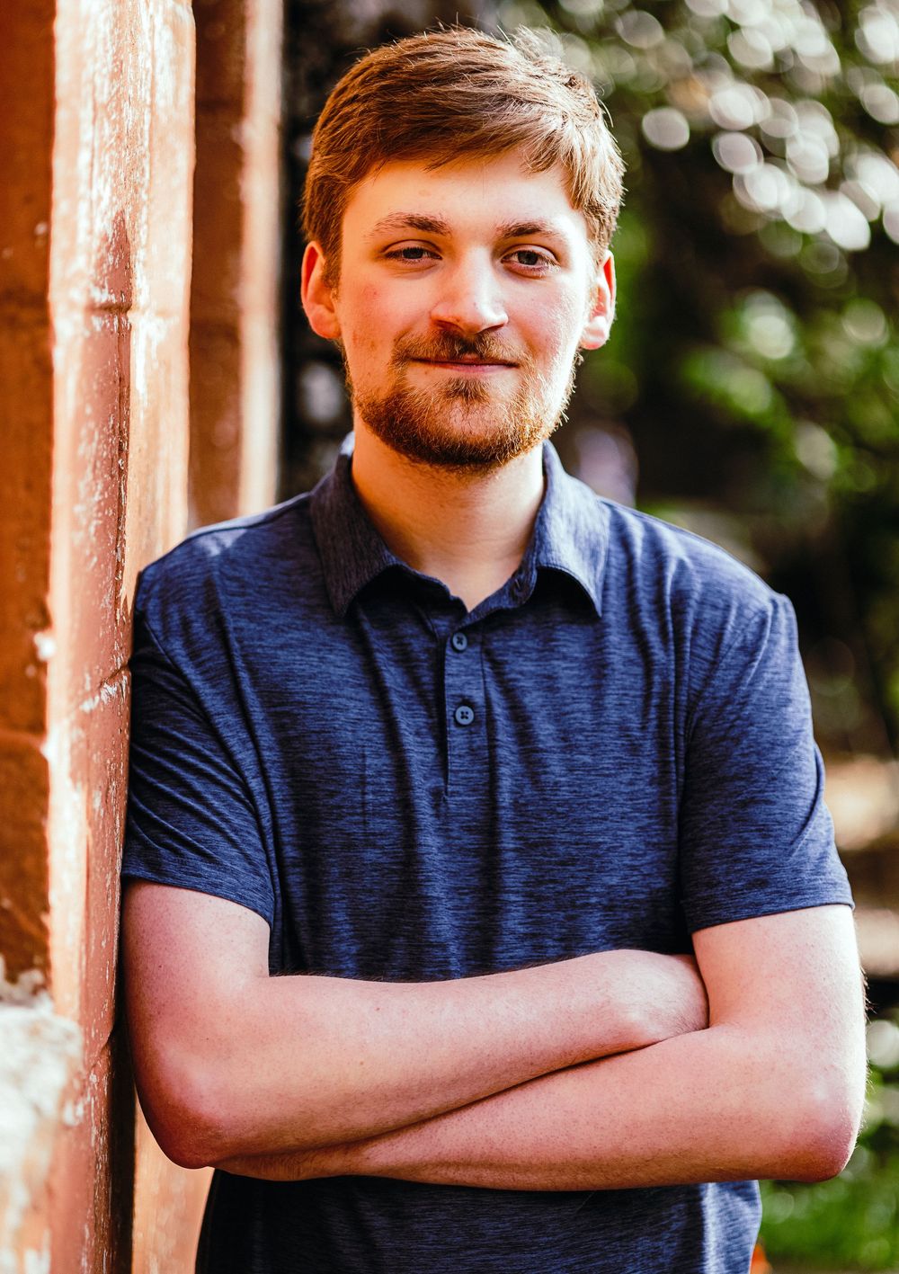 Young man with beard in blue polo shirt, arms crossed, smiling confidently while leaning against a brick wall outdoors.