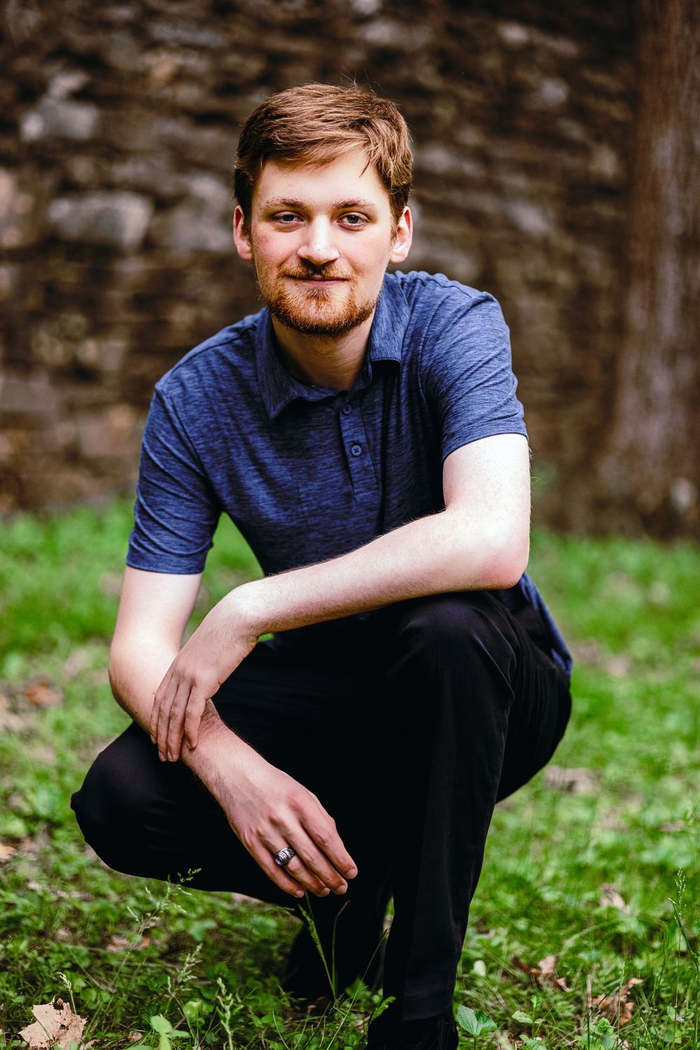 Young man with beard crouching on grass, wearing blue polo shirt and black pants, smiling outdoors near stone wall.