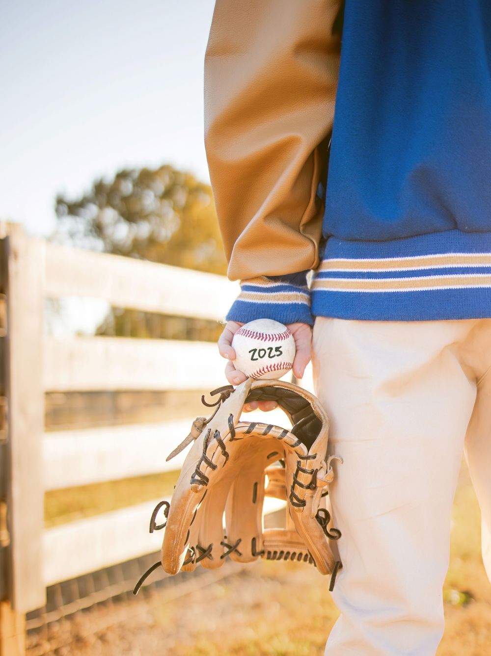 A baseball player in a blue varsity jacket holds a 2025 baseball and glove near a wooden fence outdoors.
