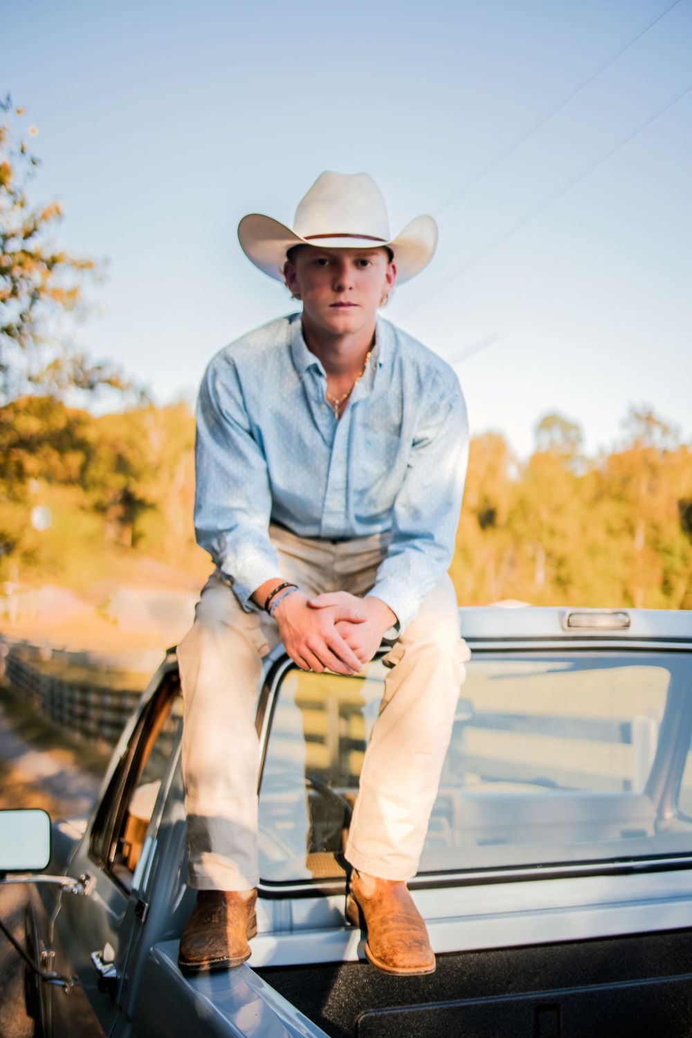 Young man in white cowboy hat and boots sits on truck hood at golden hour in rural setting.
