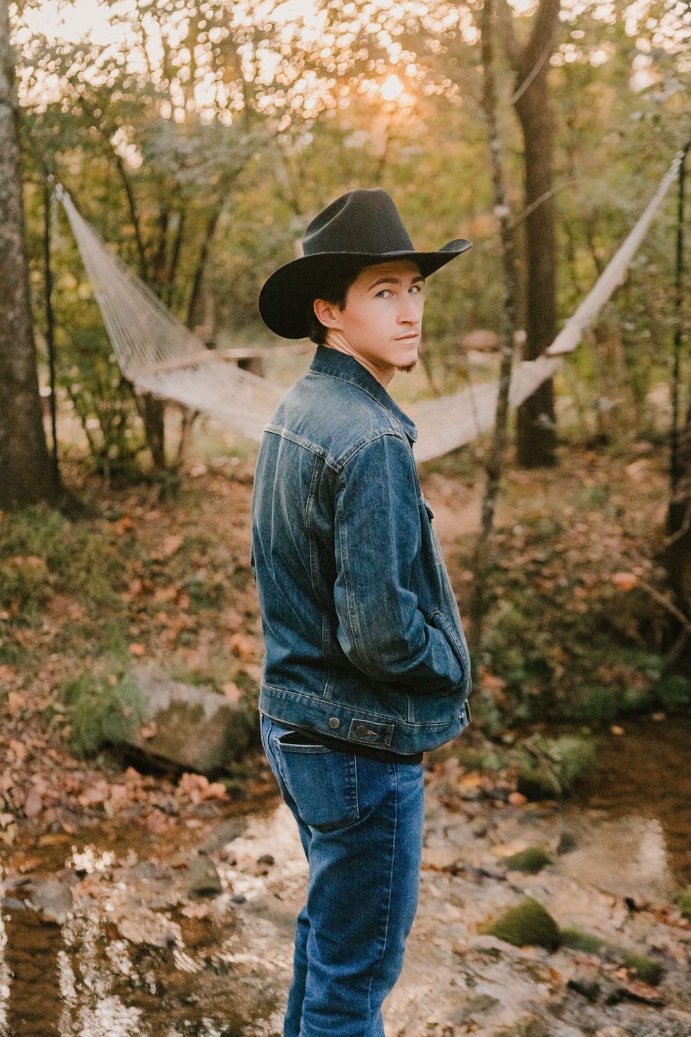 Young man in denim jacket and black cowboy hat poses outdoors near hammock and trees during golden hour.