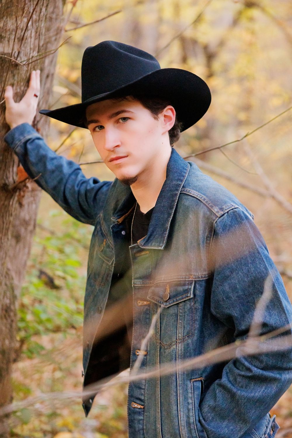 Young man in black cowboy hat and denim jacket leaning against tree in autumn forest setting.
