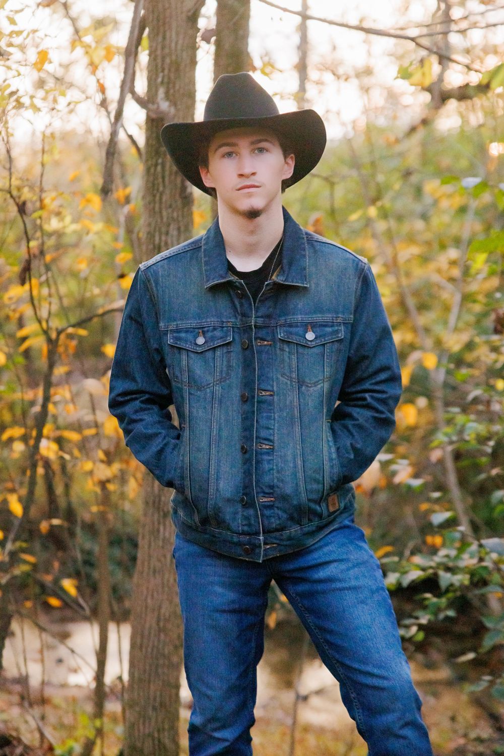 Young man in black cowboy hat and denim jacket standing in autumn forest setting.
