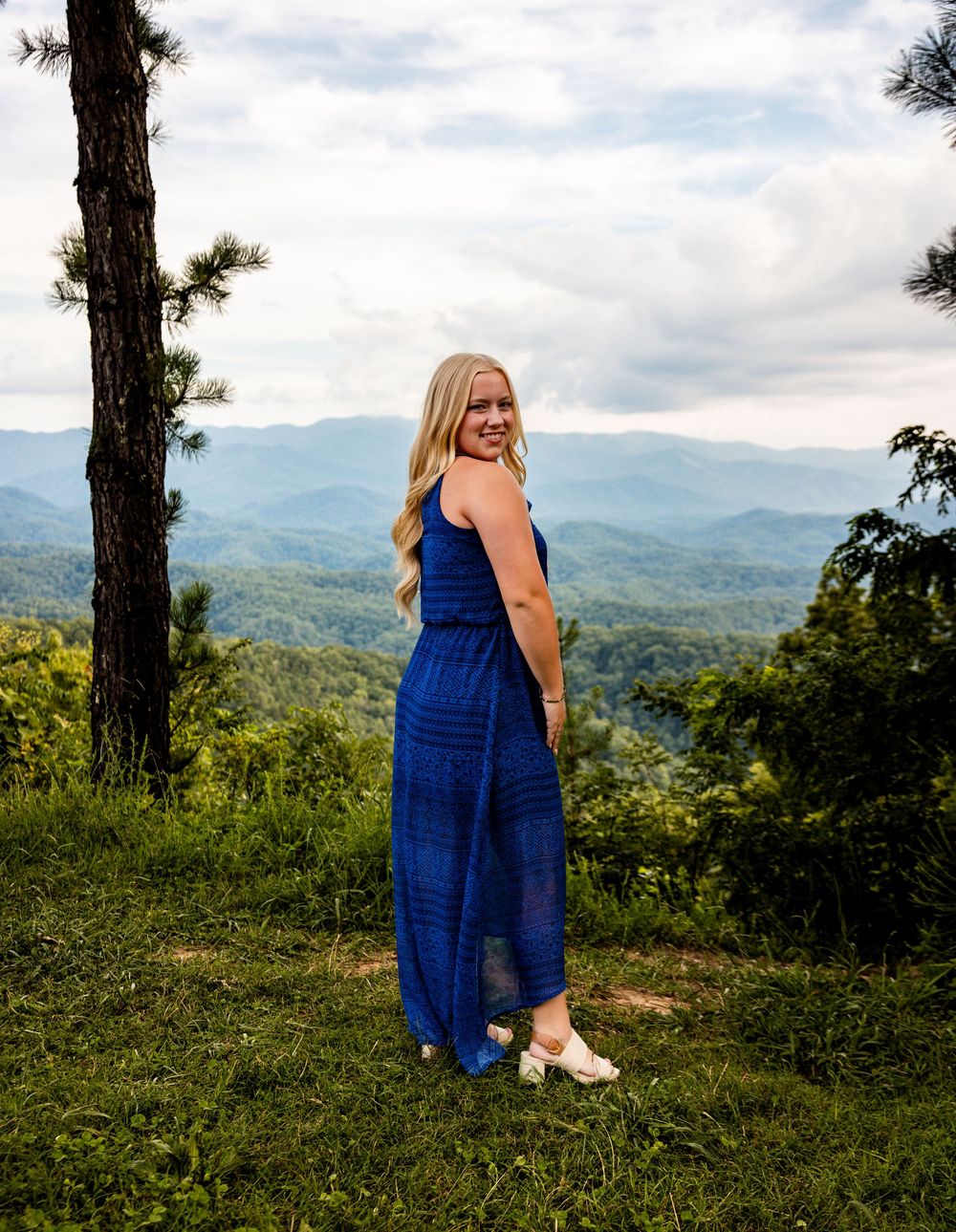 Young blonde woman in blue maxi dress posing on grassy overlook with mountain views and cloudy sky.