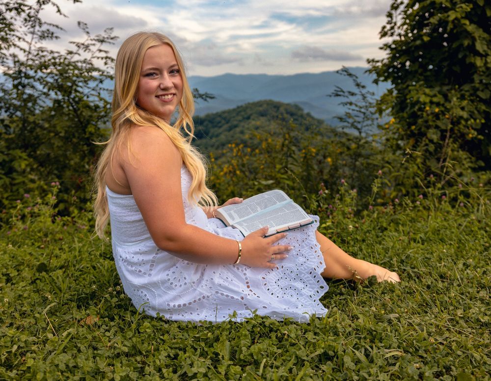 Young blonde woman in white lace dress sitting on green hillside holding a book with mountain views.
