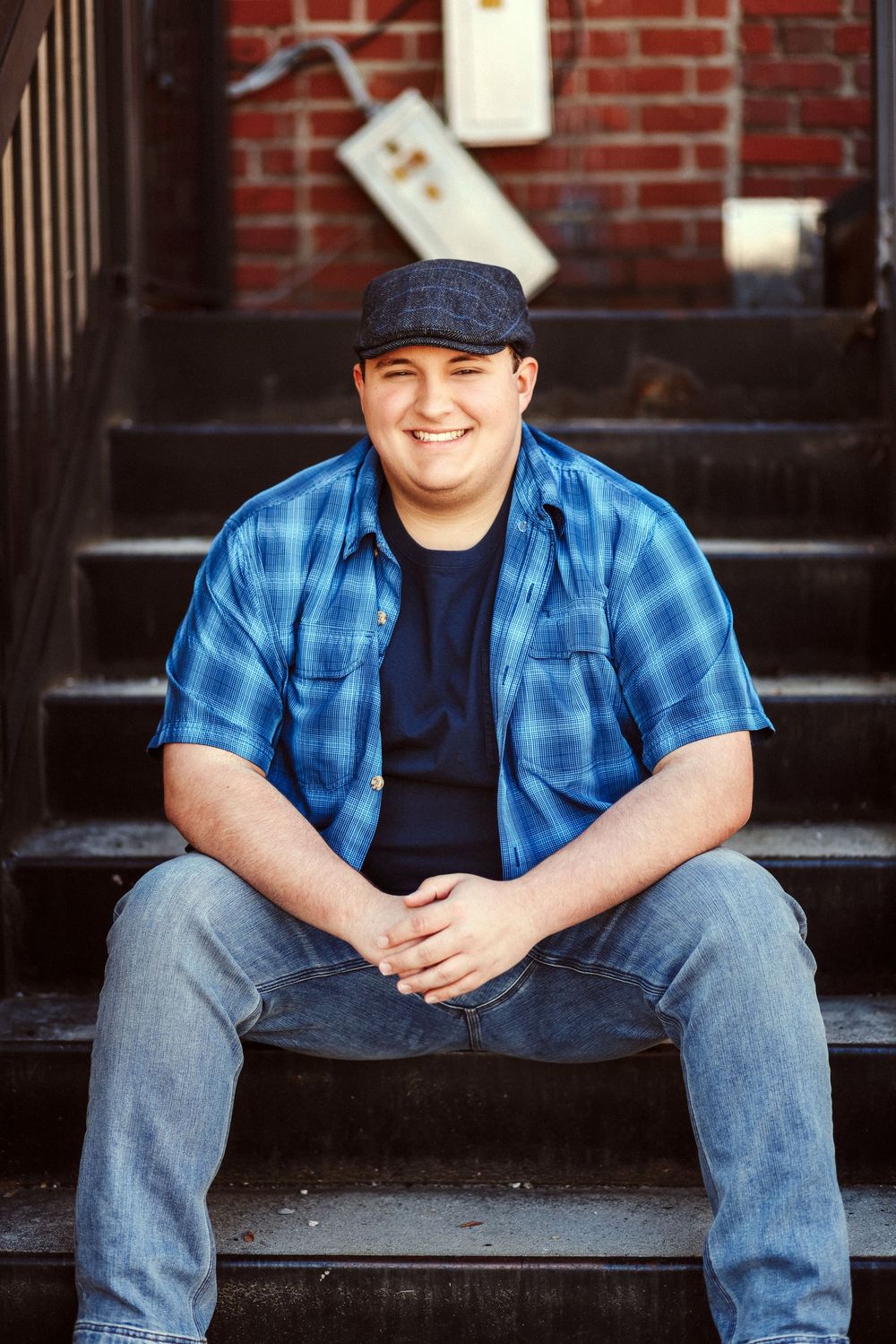 Smiling young man in blue plaid shirt and cap sitting on outdoor steps in front of brick building.
