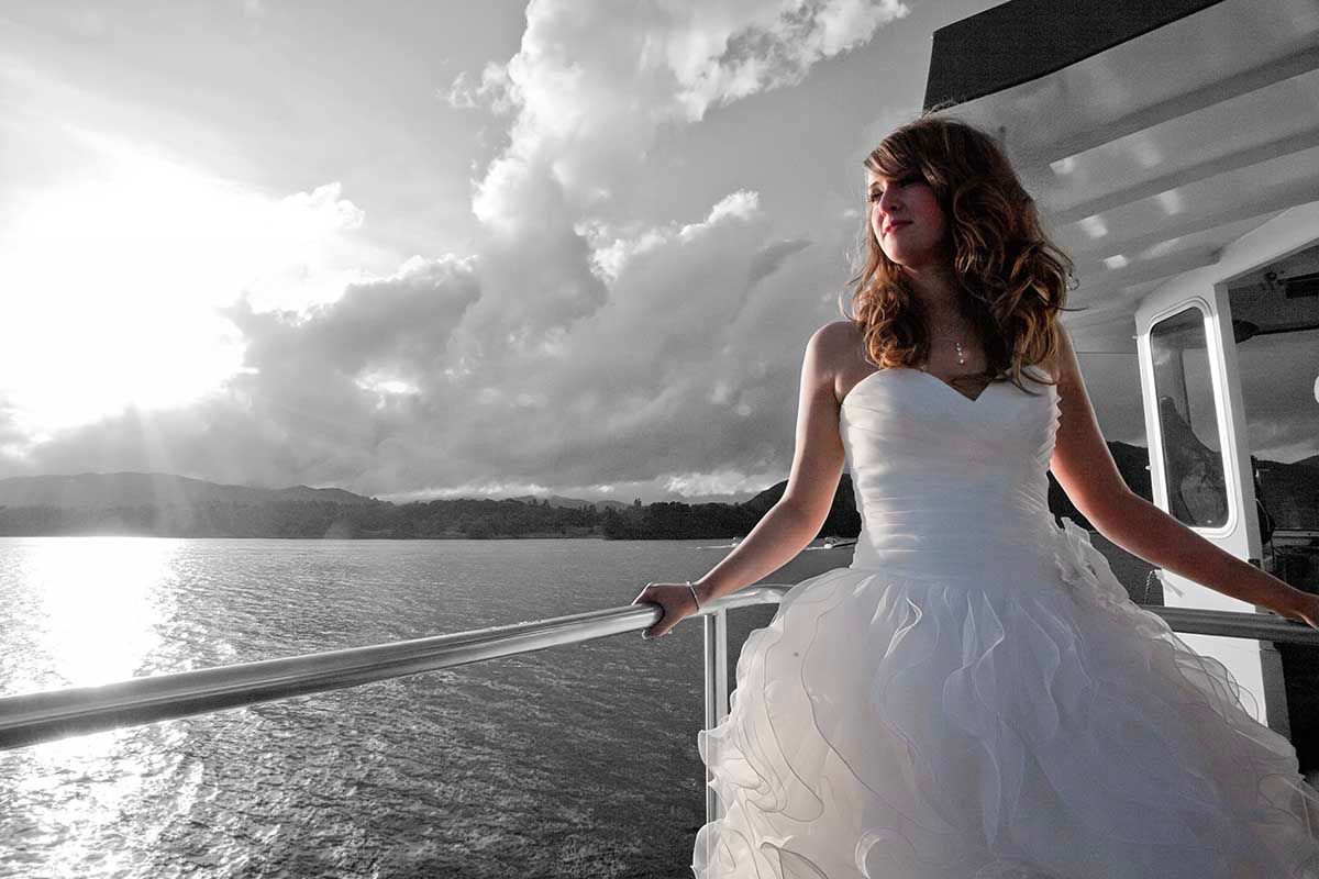 Bride in white wedding gown standing on cruise ship deck, gazing at dramatic cloudy sky over water.