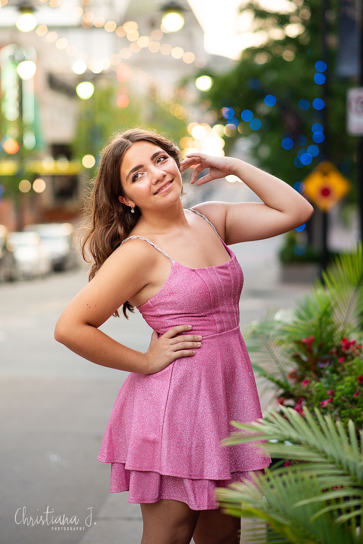Young woman in pink sparkly dress posing on a vibrant city street at dusk.