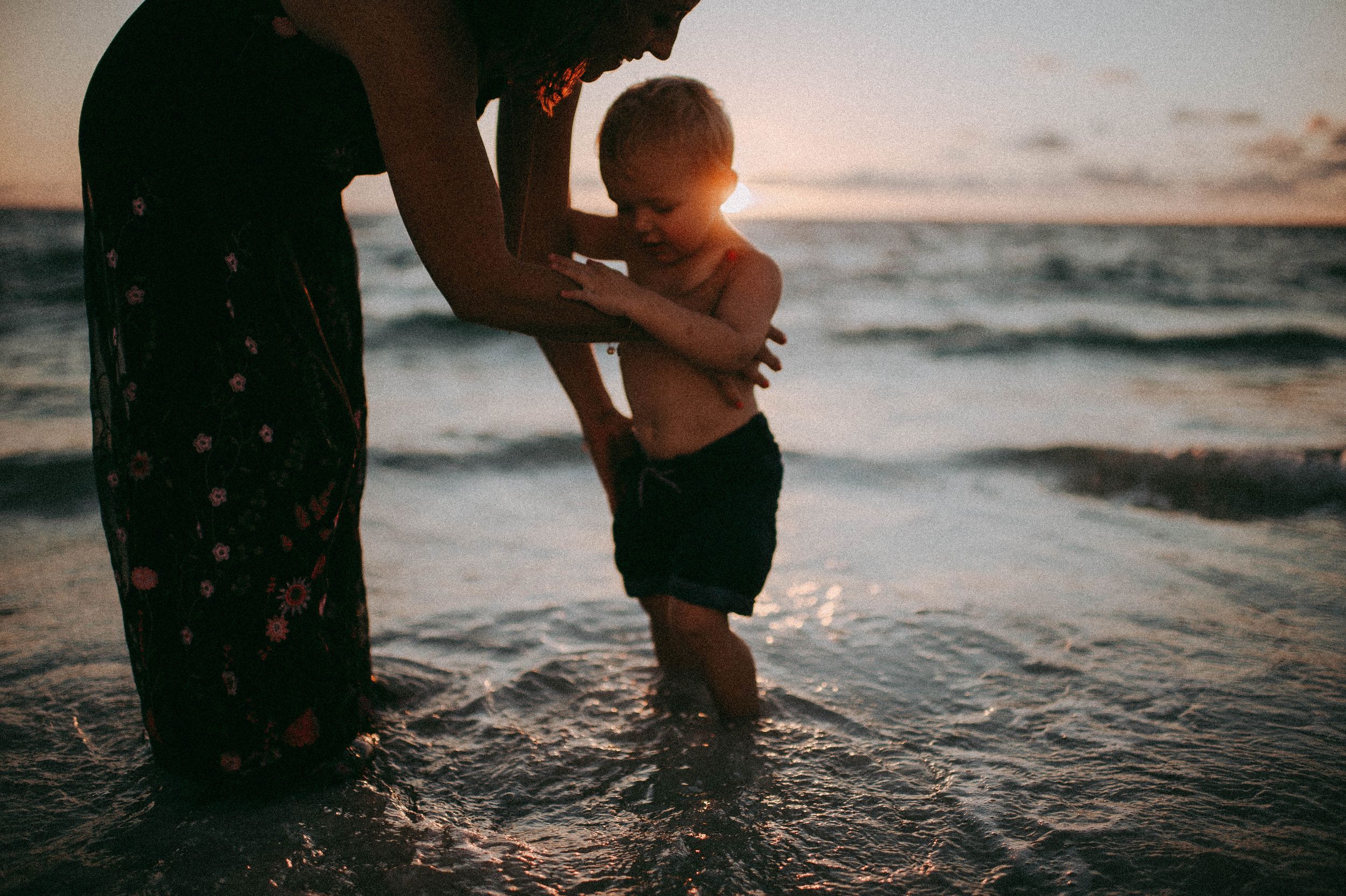 Mother holding toddler at sunset beach, waves gently washing over their feet in golden evening light.