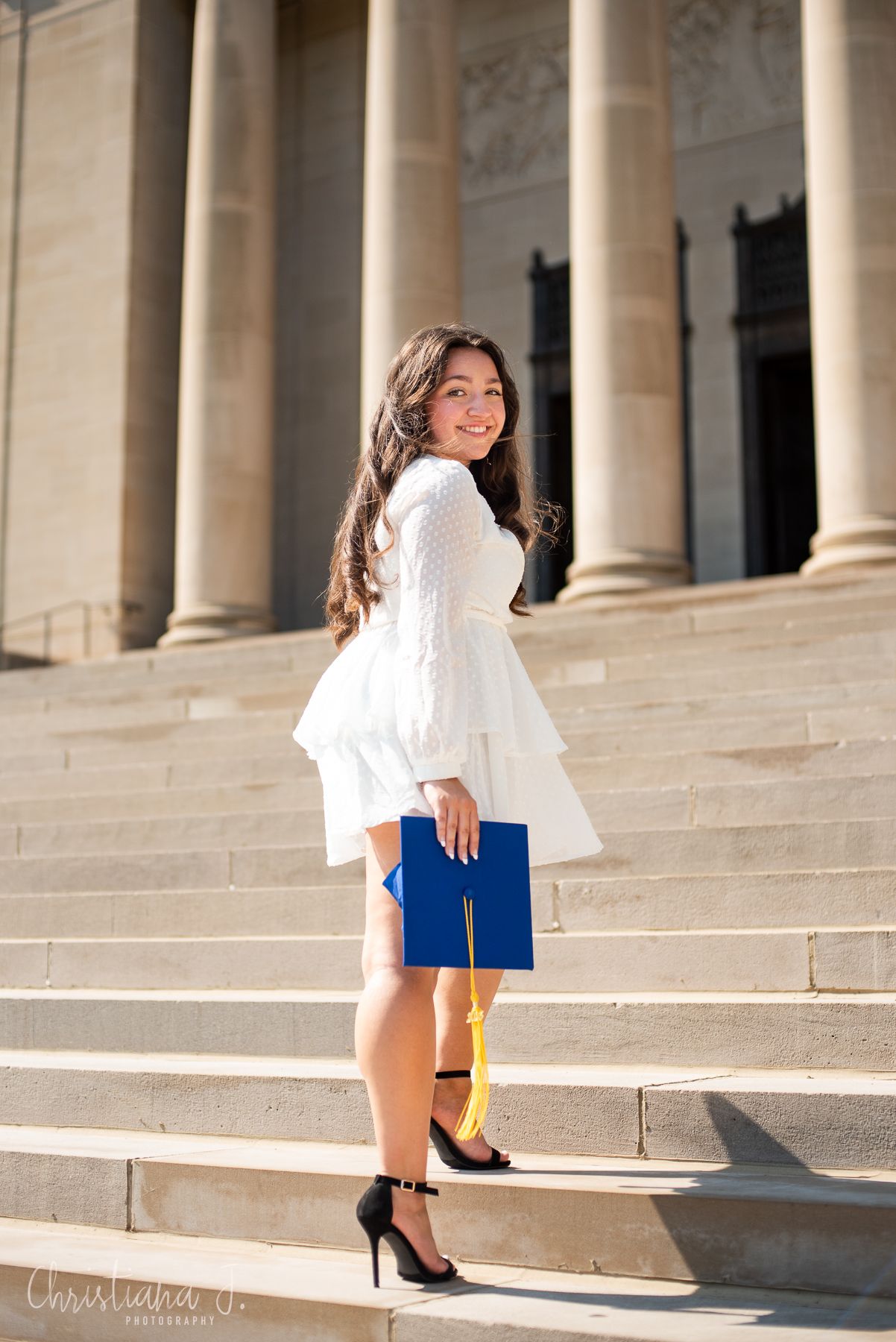 Graduate in white dress holding blue cap on steps of a grand columned building.