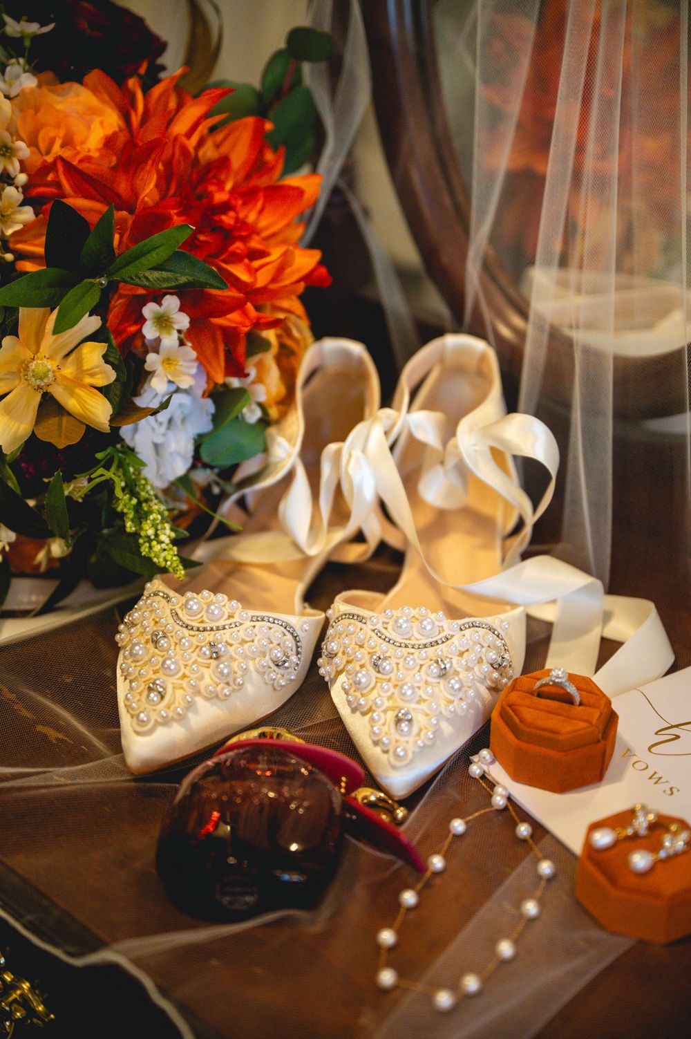 Elegant bridal flat shoes adorned with pearls and crystals beside a colorful bouquet, orange velvet ring box, and sheer veil.