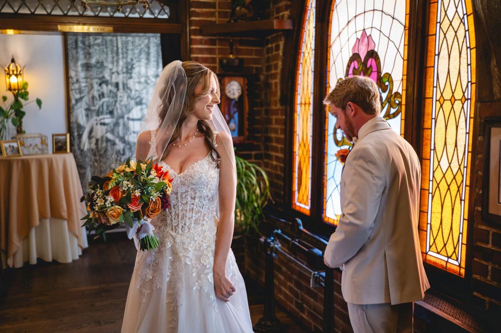 Bride in lace gown holds bouquet facing groom in tan suit before ornate stained glass window at wedding venue.