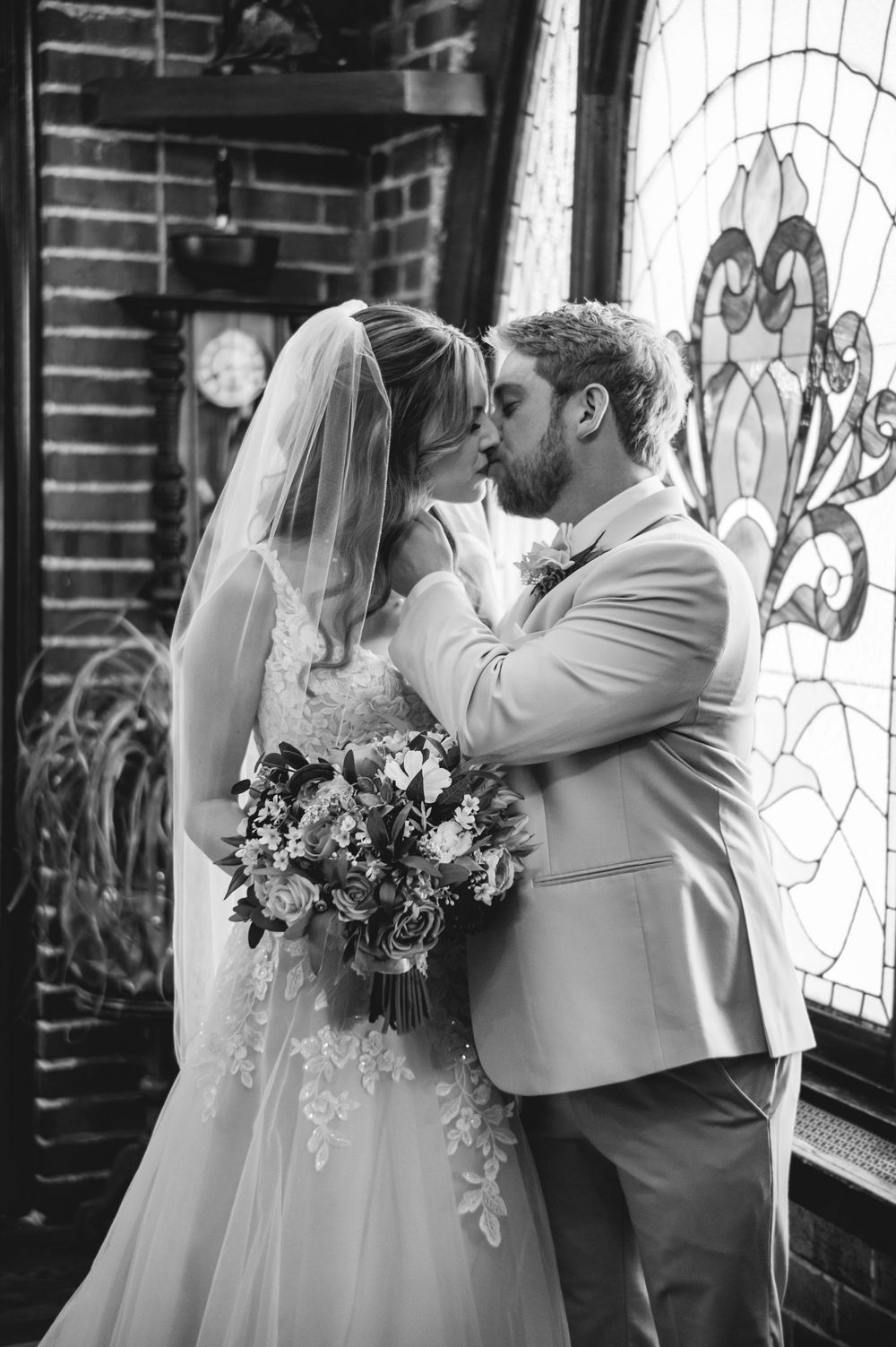 Black and white wedding photo of bride and groom sharing a kiss near a stained glass window.