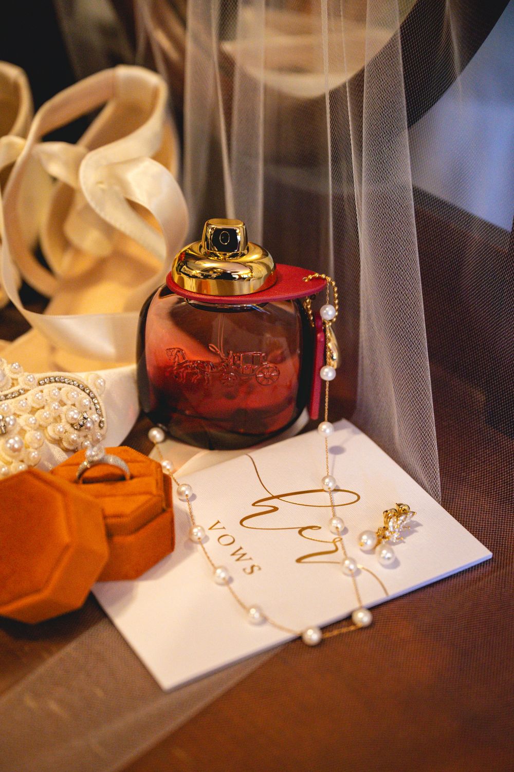 Bridal flat lay with red perfume bottle, vow book, pearl necklace, ring box, and wedding veil on dark surface.