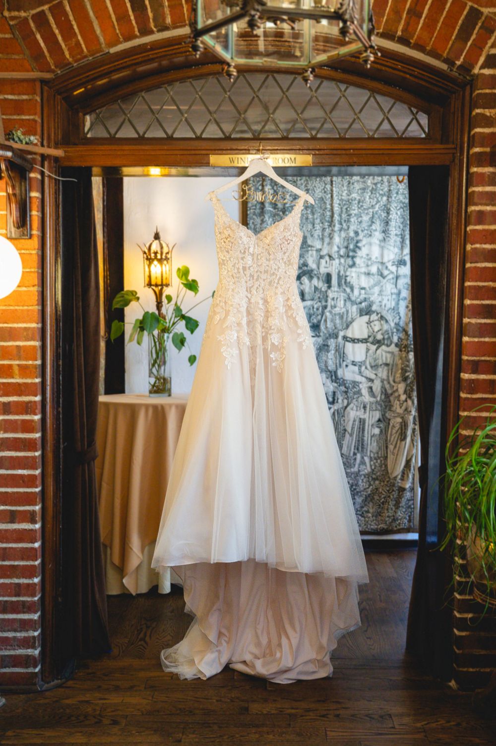 Elegant lace wedding dress hanging in a rustic brick doorway with warm ambient lighting.