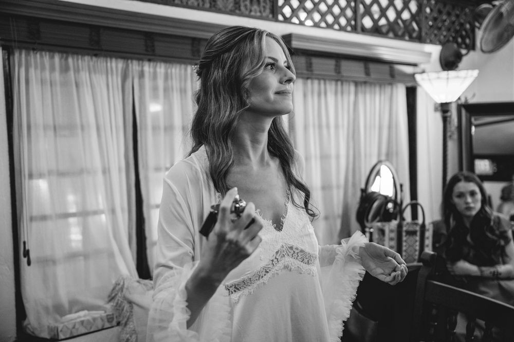 Bride in white lace robe applying perfume before wedding ceremony in black and white photo.