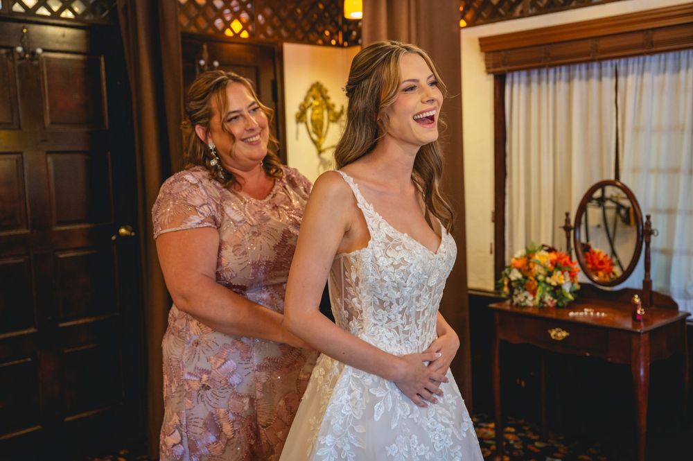 Bride laughing joyfully as woman helps her into elegant white lace wedding gown in a warmly decorated bridal suite.