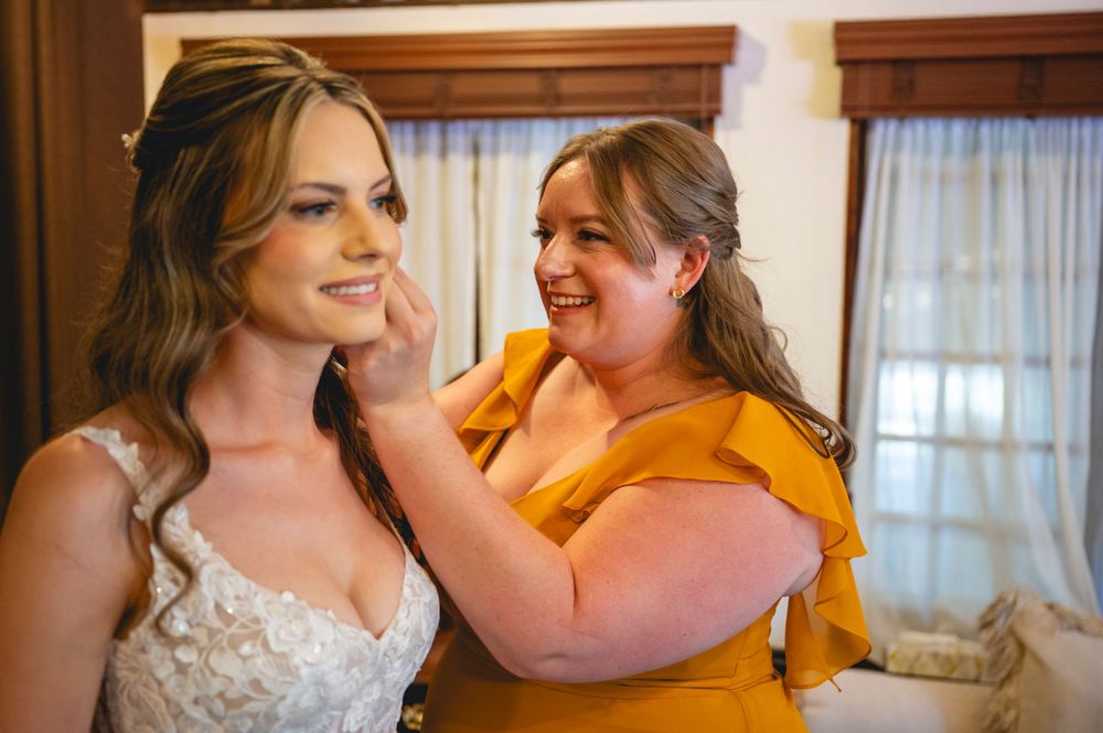 Bride in lace gown smiles as woman in yellow dress touches her face during wedding preparations.