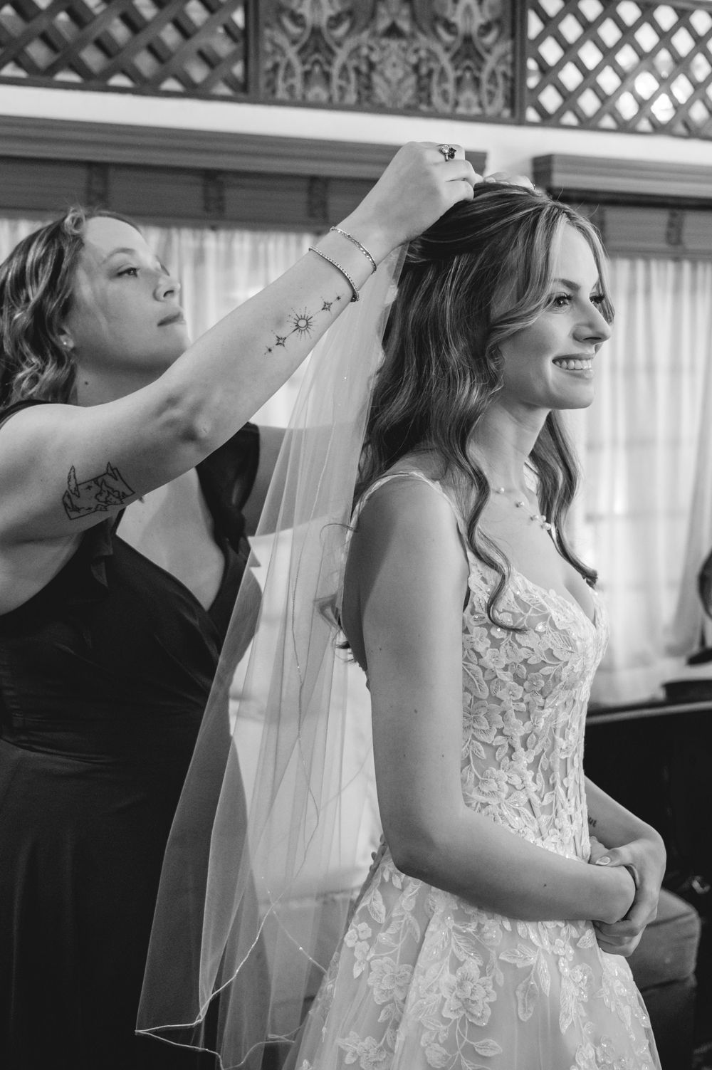 Bridesmaid adjusting veil on smiling bride in lace wedding dress, black and white photo.