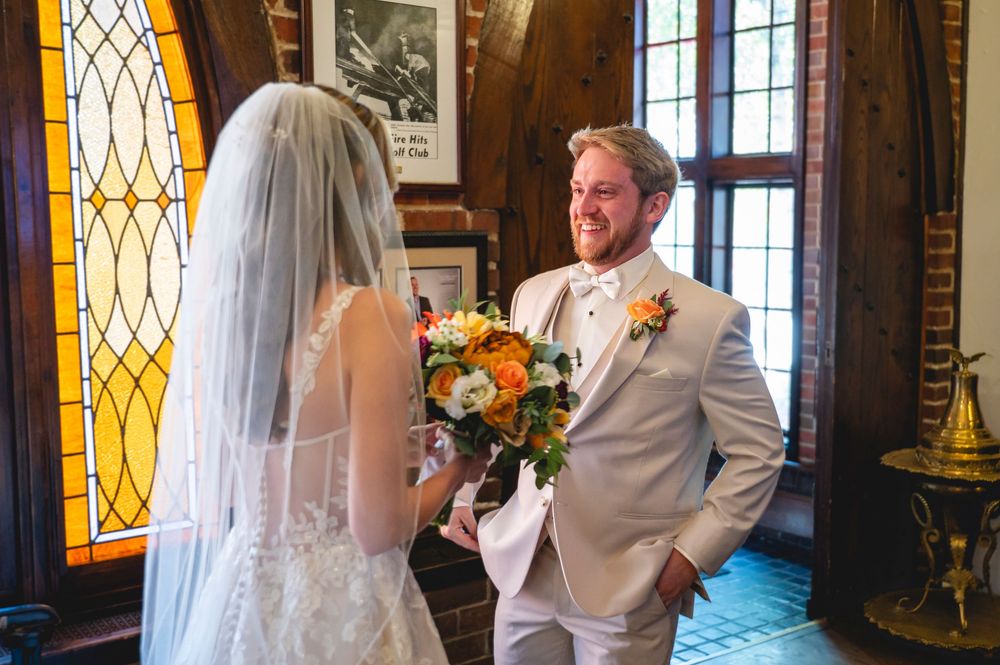 Groom in white suit smiles at bride holding colorful bouquet during wedding first look in elegant venue.