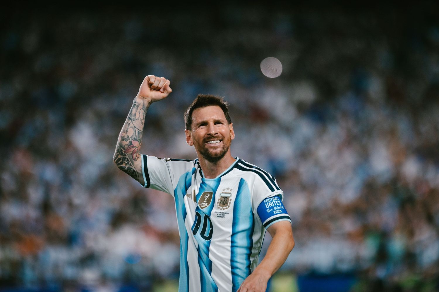 Soccer player in Argentina jersey celebrates with raised fist, tattoos visible, stadium crowd in background.
