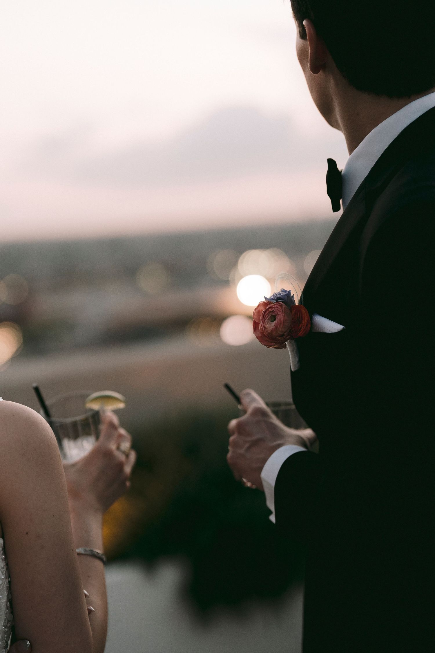 Couple in formal wedding attire holding cocktails at sunset rooftop, groom wearing black tuxedo with red boutonniere.