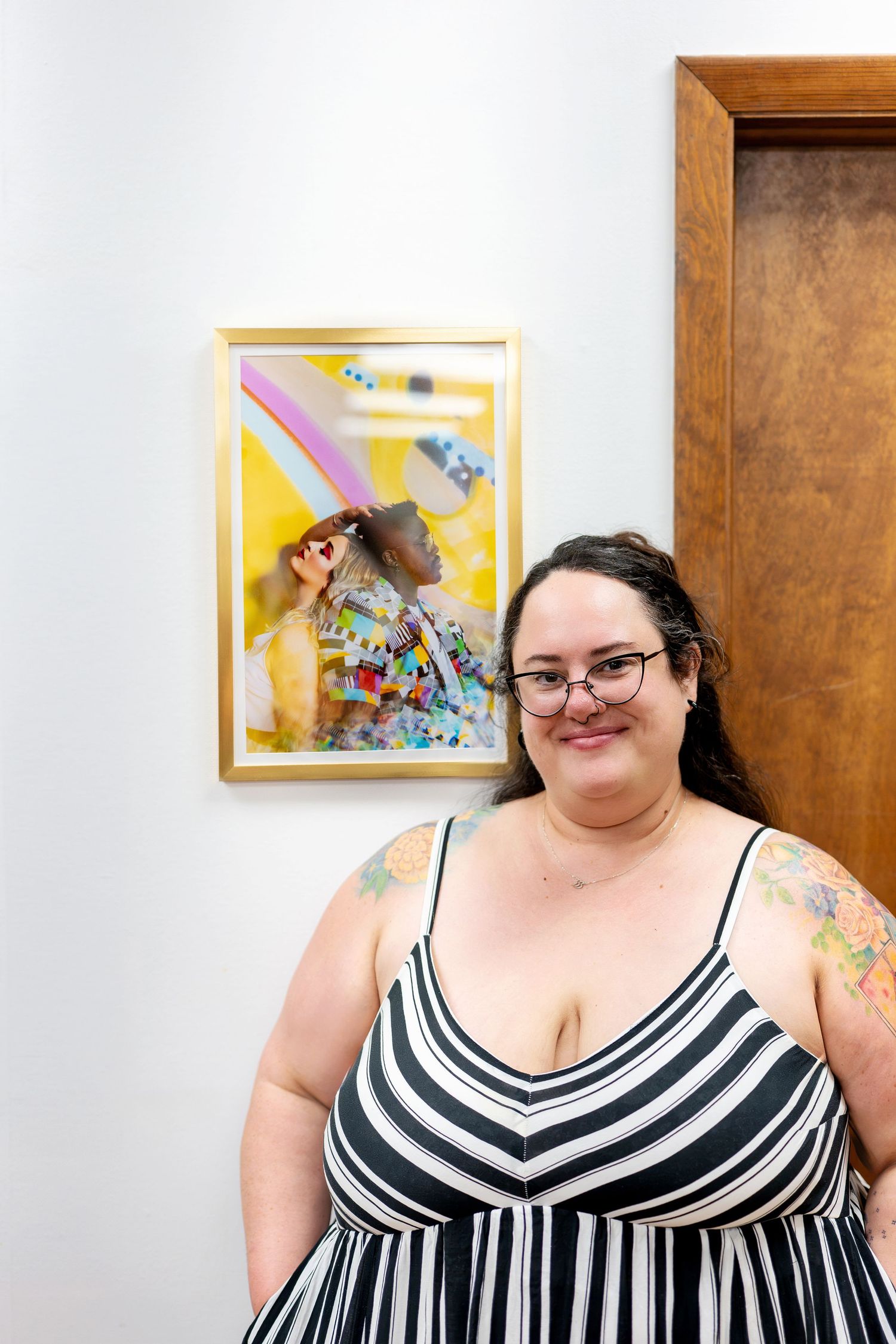 Plus-size woman with tattoos and glasses in striped dress smiling in front of colorful framed artwork.