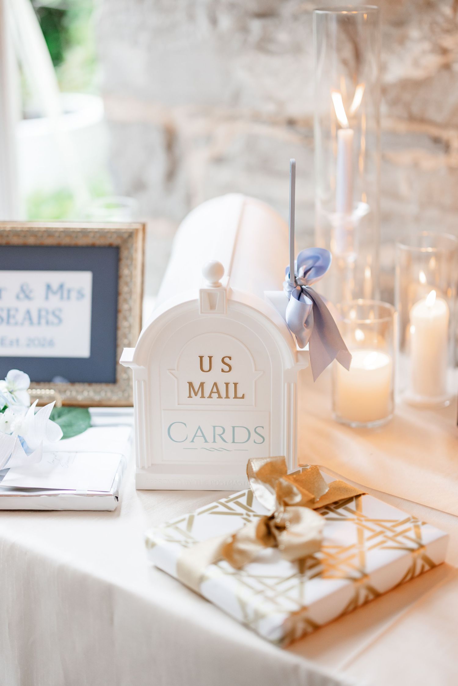 White mailbox card holder with blue ribbon beside candles and gifts on wedding gift table.