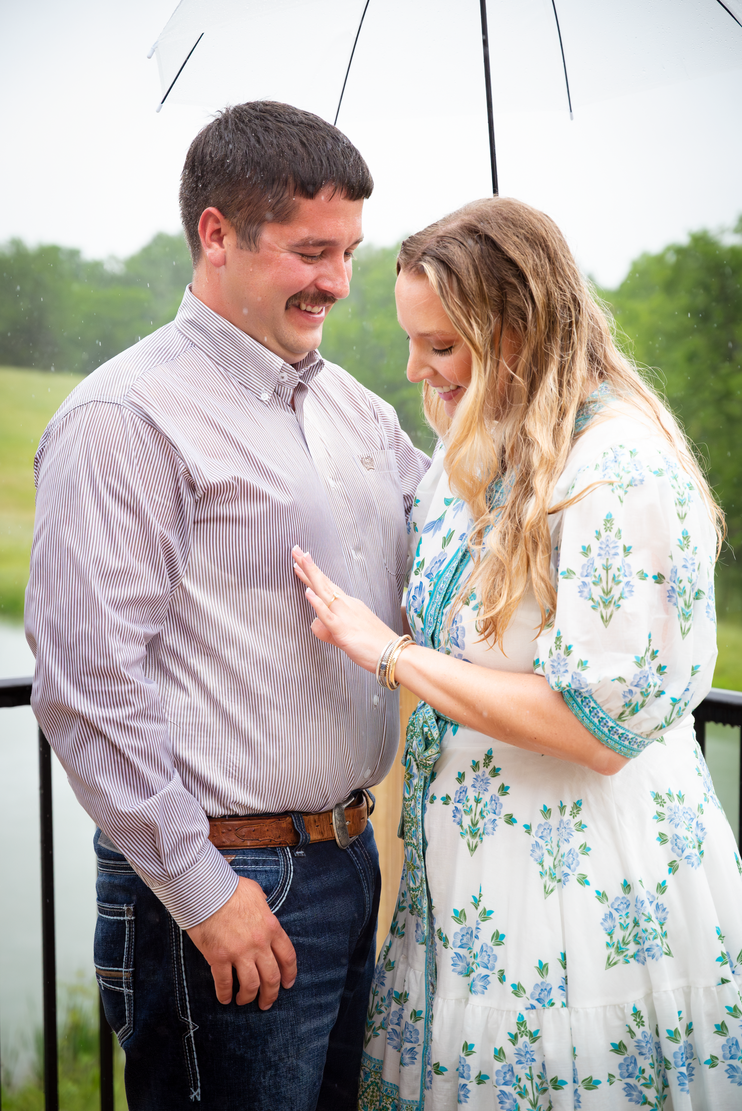 A couple shares a romantic moment under a clear umbrella in the rain, smiling at each other outdoors.