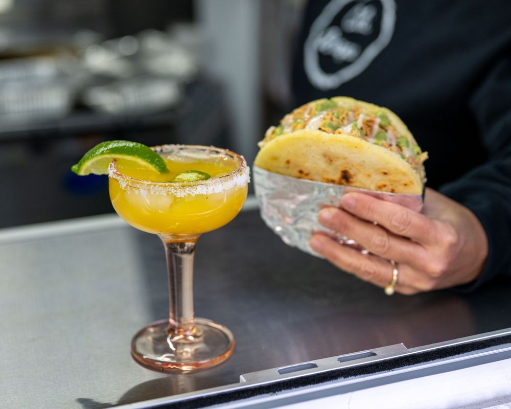 A golden margarita with salted rim and lime garnish beside a hand holding a taco at a food stand.