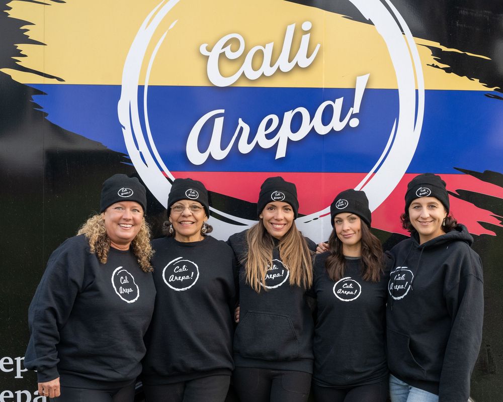 Five women in black Cali Arepa hoodies pose proudly in front of a colorful Colombian-themed food truck logo.