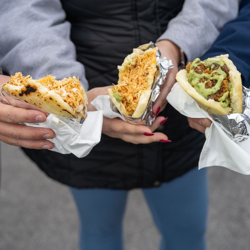 Three people holding arepas and tacos wrapped in foil and paper at an outdoor food market.