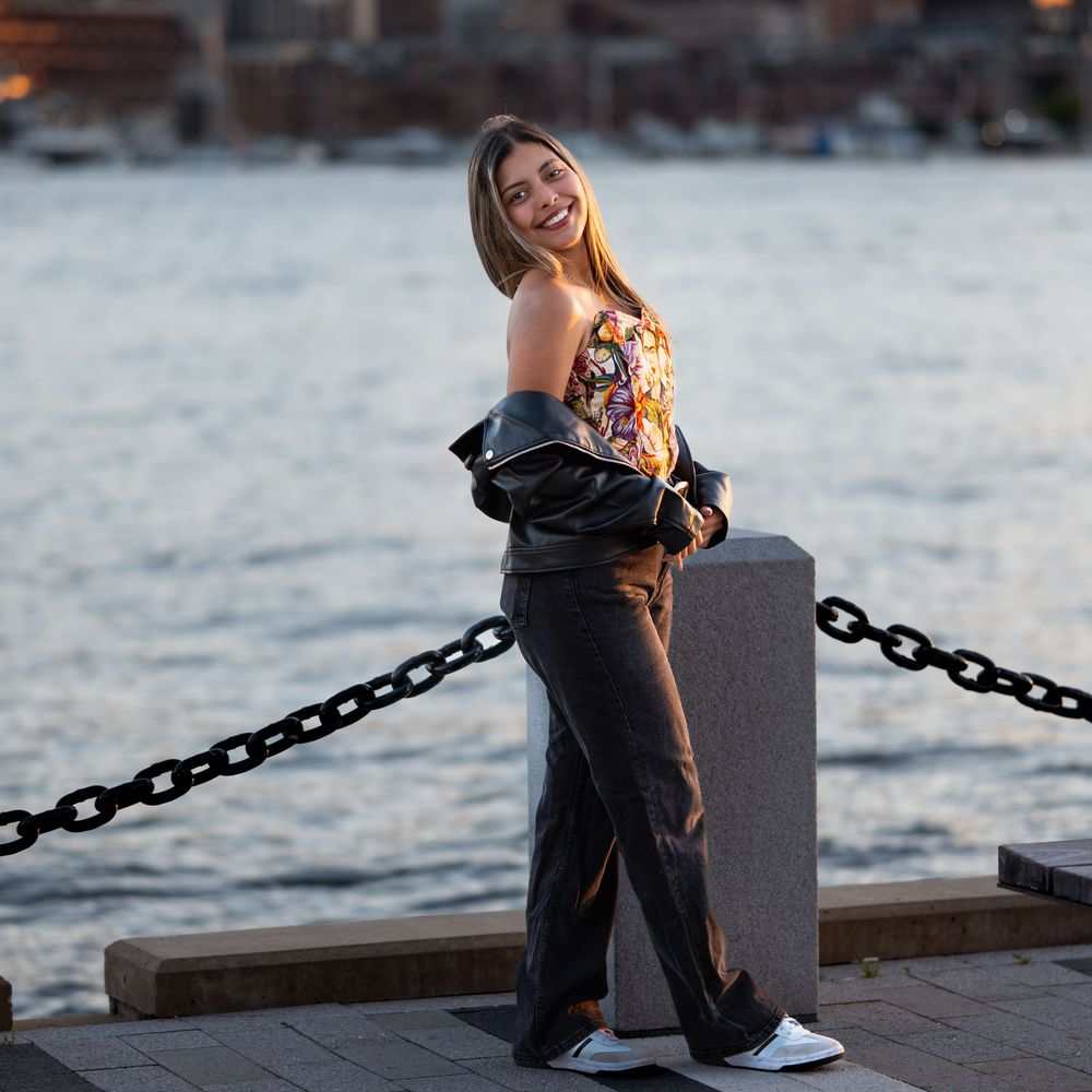 Woman in floral top and black pants smiling on a waterfront promenade at dusk.