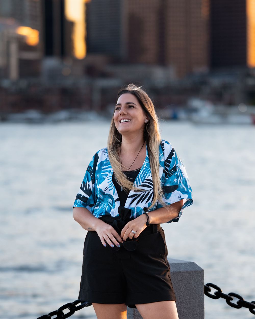 Woman in floral blue shirt and black shorts smiling by waterfront with city skyline at golden hour.