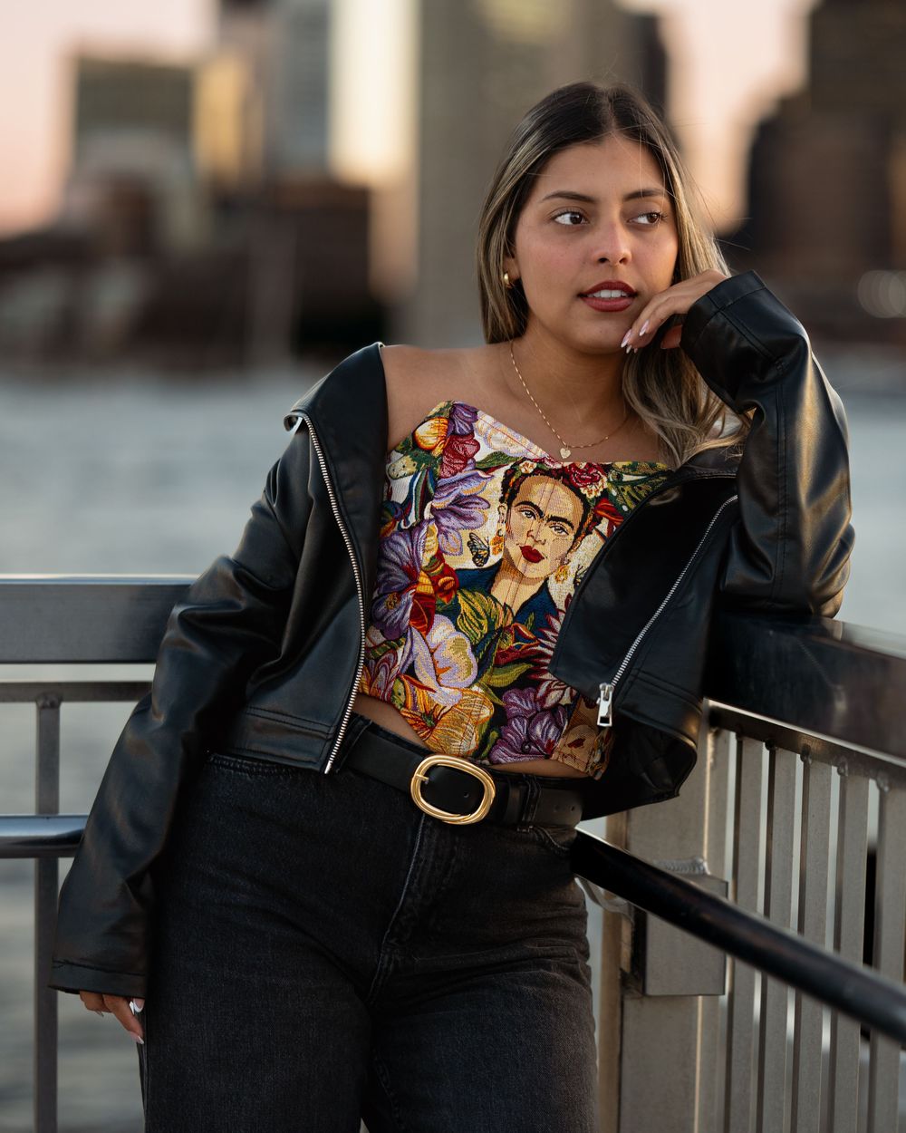 Woman in black leather jacket and Frida Kahlo top poses by waterfront railing with city skyline at sunset.
