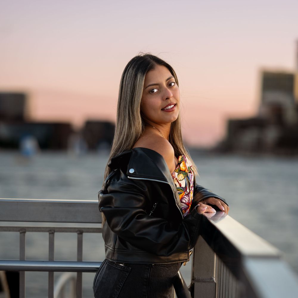 Young woman in black leather jacket poses by waterfront railing at sunset with city skyline in background.