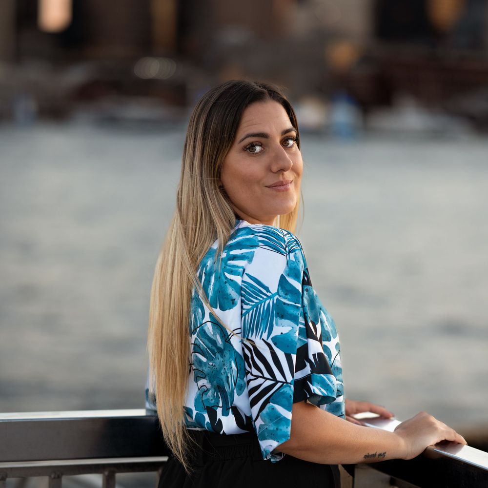 A young woman with long blonde hair wearing a tropical print blouse poses outdoors near a railing with a blurred urban background.
