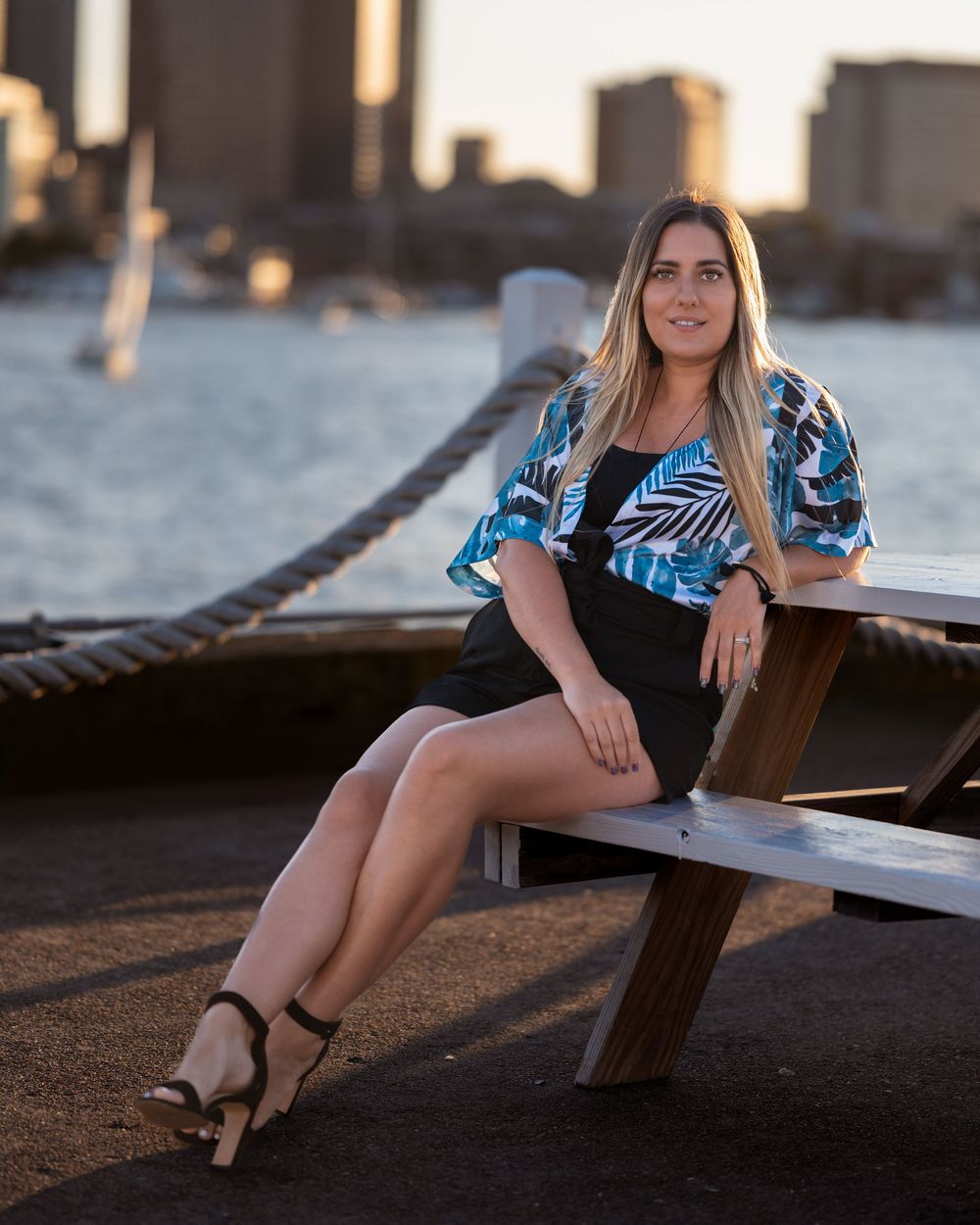 Woman in tropical print jacket sits at waterfront picnic table with city skyline at sunset.