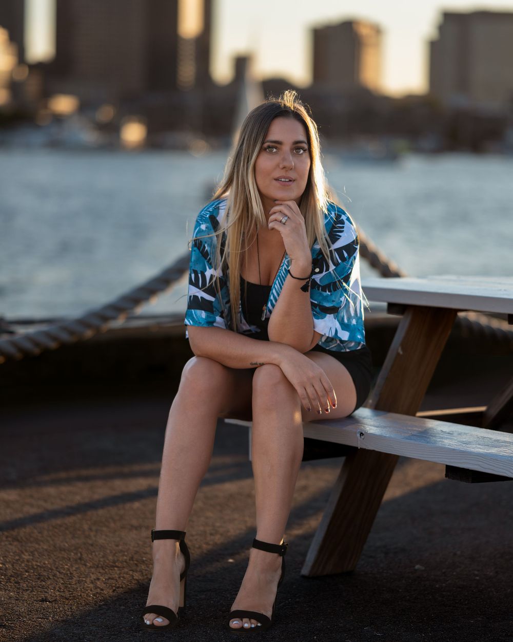 Woman in floral kimono sitting on waterfront bench with city skyline at golden hour.
