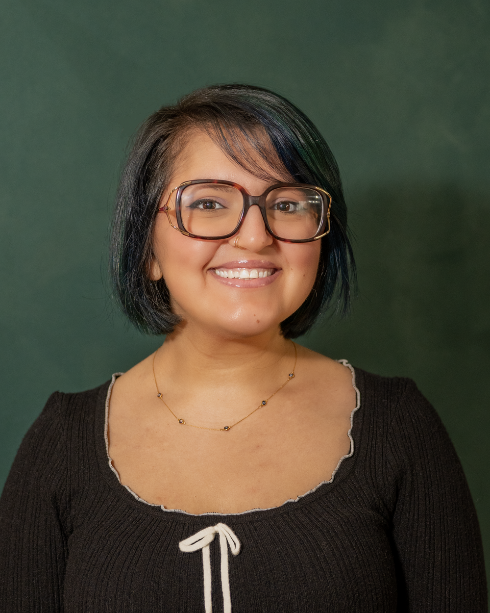 Young woman with short dark hair, glasses, and a black top smiles warmly against a green background.
