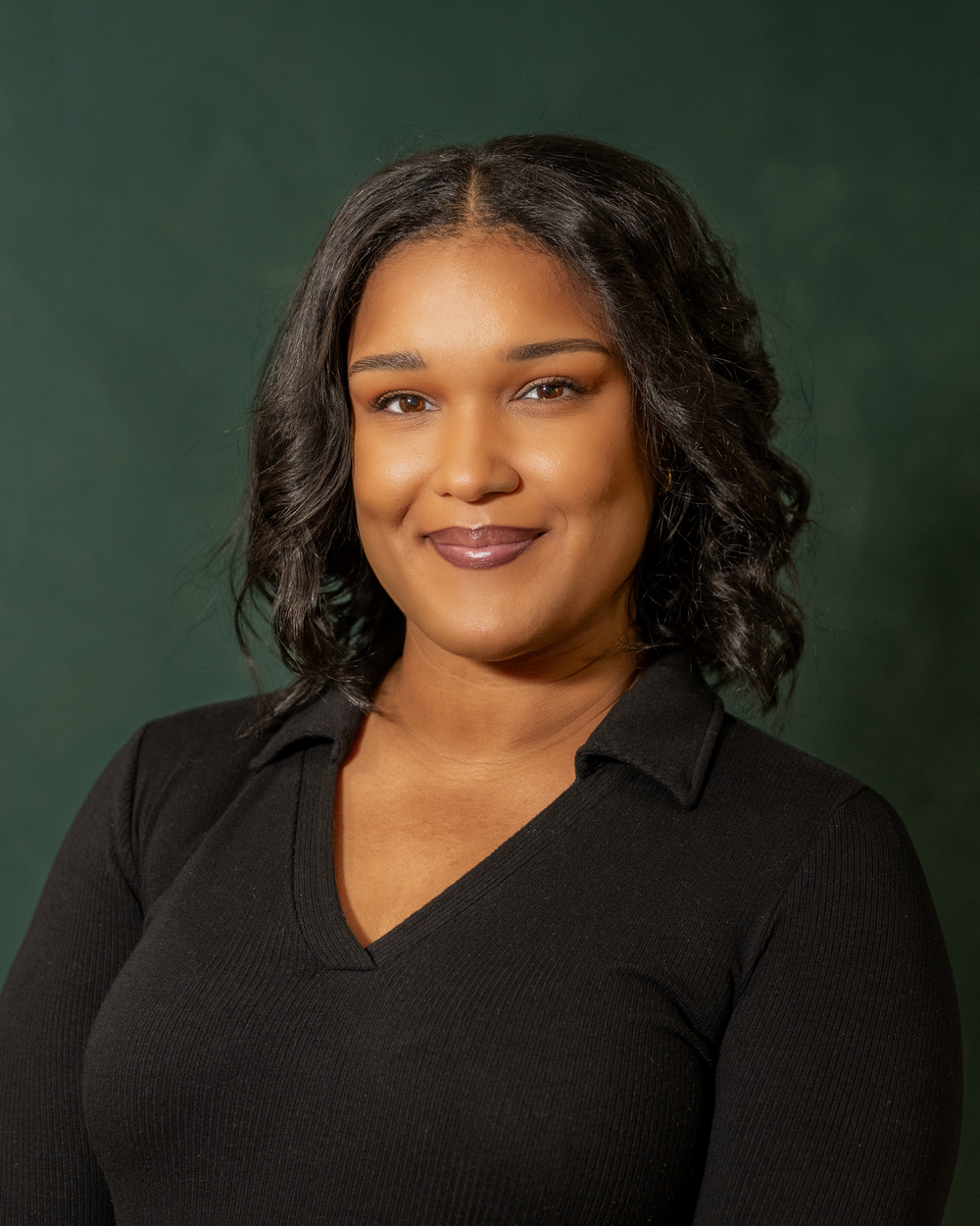 Professional headshot of a smiling woman in a black v-neck top against a dark green background.