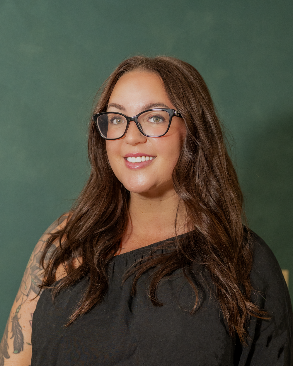 A smiling woman with long brown hair and black glasses wearing a dark top with a tattoo on her arm against a green background.