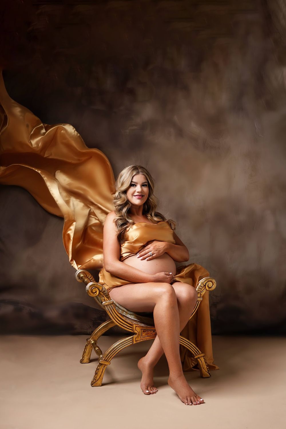A smiling pregnant woman in gold satin fabric sits on an ornate chair with flowing fabric in a studio.