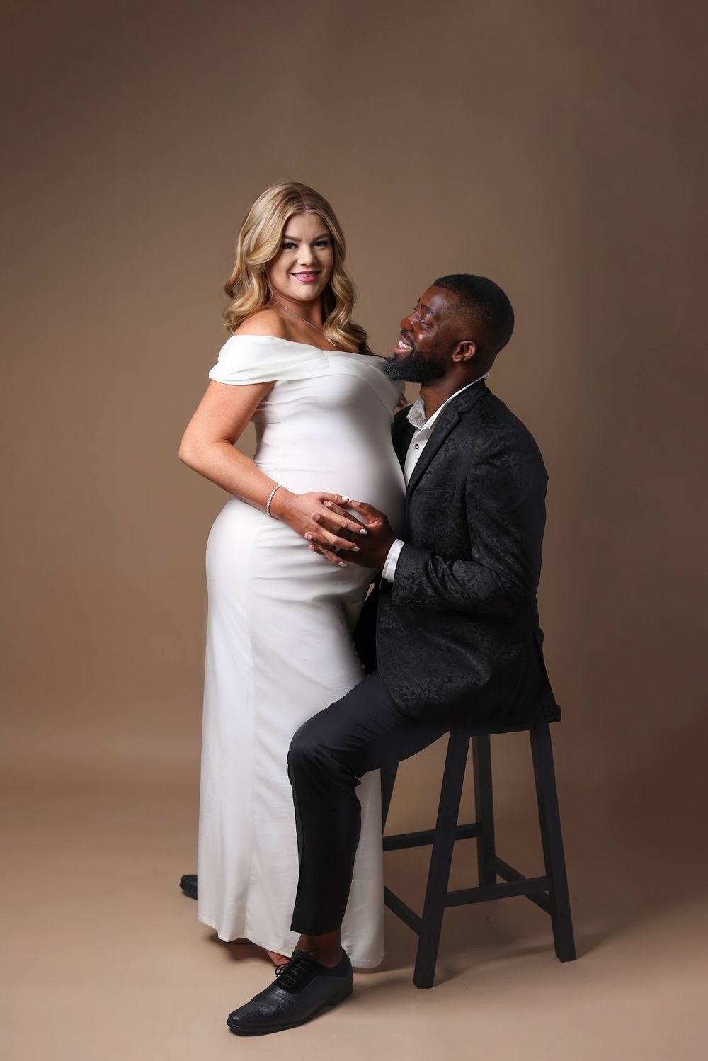 A pregnant woman in a white off-shoulder gown stands beside her seated smiling partner in a studio portrait.